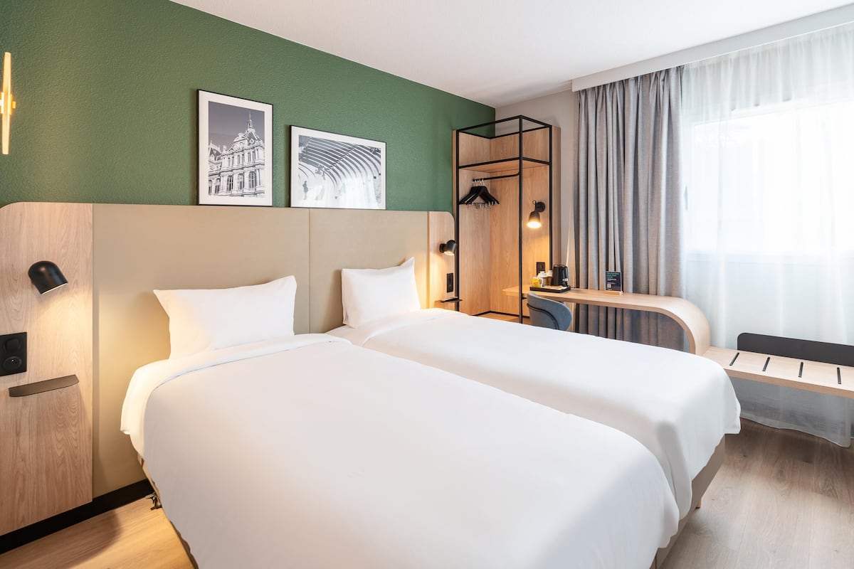 A hotel room with two beds, a desk, a chair, a mirror, and a window with curtains.