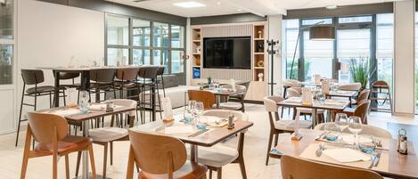 A modern dining area with multiple tables and chairs, a large flat-screen TV, and a view of the outdoors through large windows.