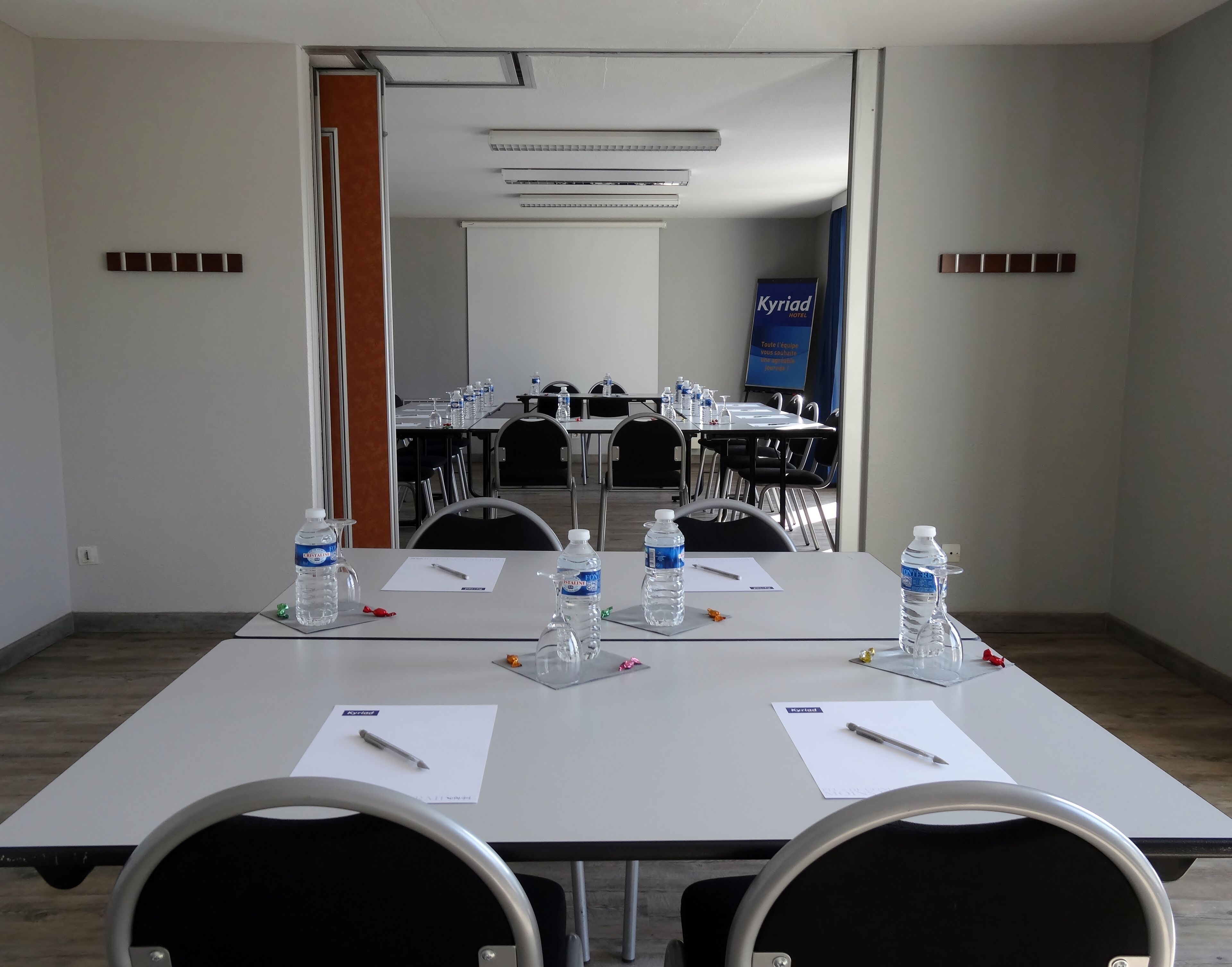 Una sala de conferencias con mesas, sillas, botellas de agua y un cartel para Kyriad Hotel.