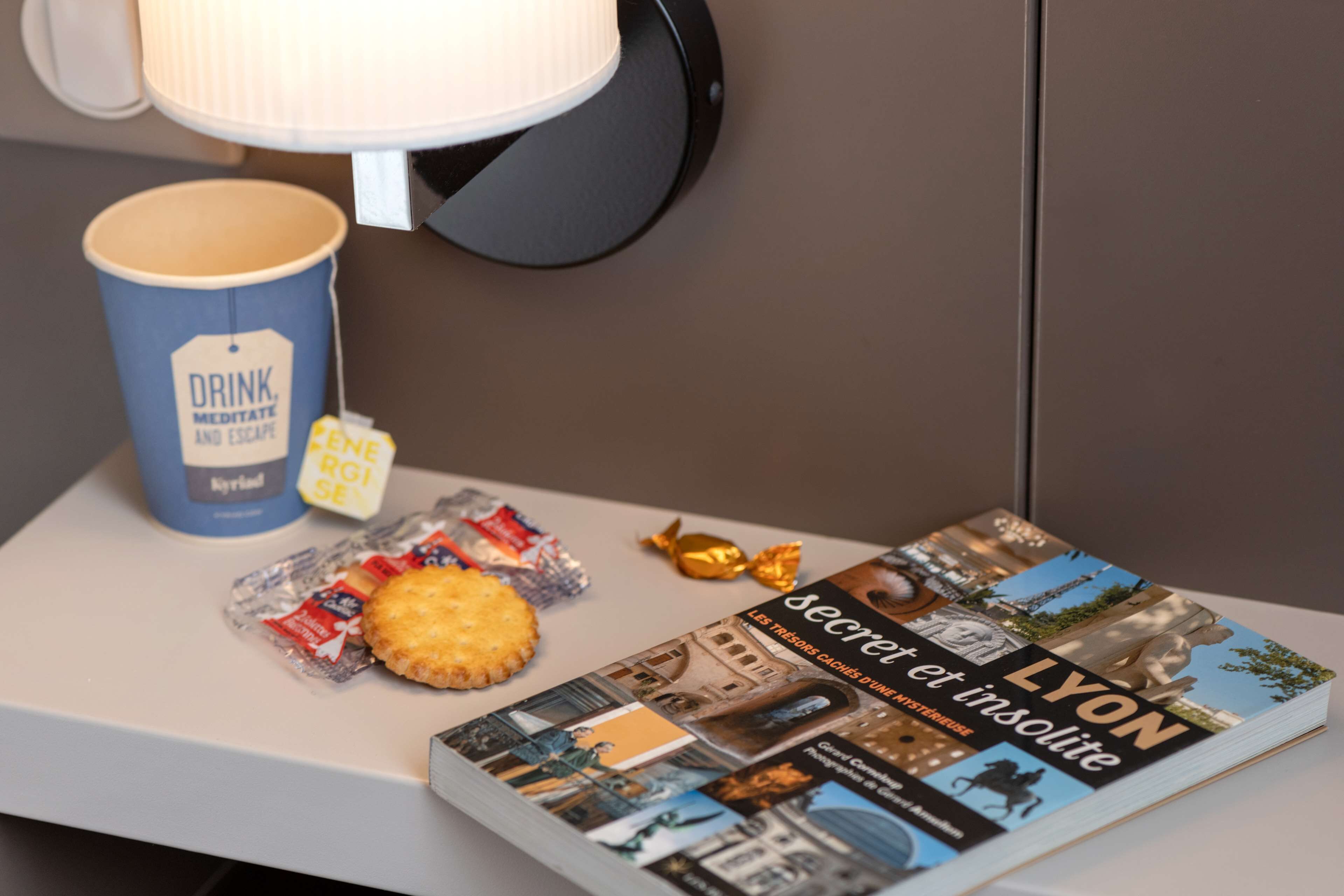 A table with a book titled 'Secret et insolite Lyon', a mug with 'DRINK, MEDITATE AND ESCAPE' text, a biscuit, and a tea bag.