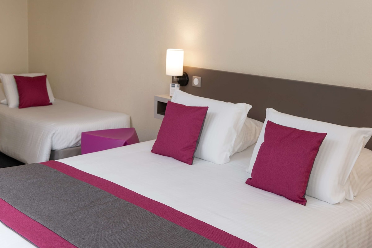 A hotel room with two single beds, each with white linens and a single pink pillow. There is a large bed with white linens and multiple pink pillows. A nightstand with a lamp is visible between the beds.