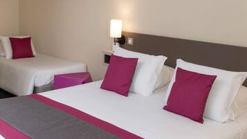 A hotel room with two single beds, each with white linens and a single pink pillow. There is a large bed with white linens and multiple pink pillows. A nightstand with a lamp is visible between the beds.