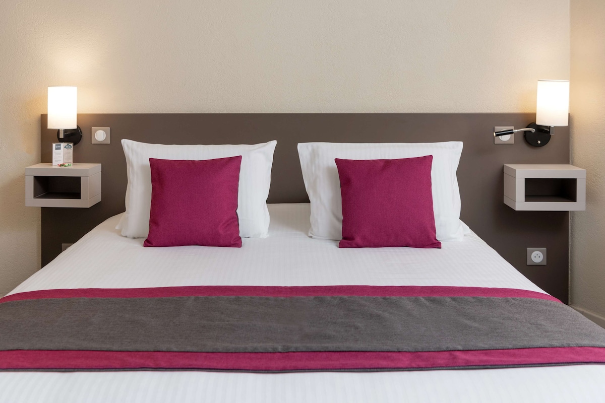 A neatly made bed with white and pink pillows, a grey and pink bedspread, and two bedside tables with wall-mounted lamps.