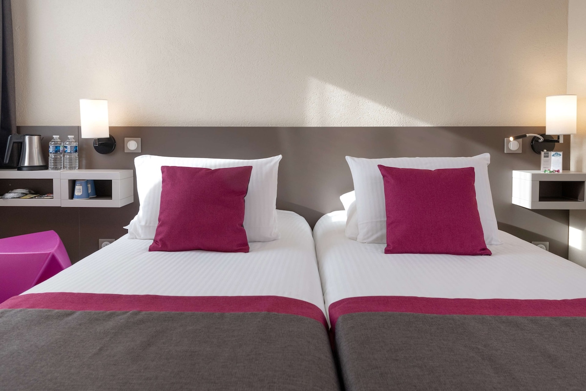 A hotel room with two beds, each with white linens and a single pink pillow. There are two bedside tables with lamps, a small shelf with toiletries, and a pink chair.