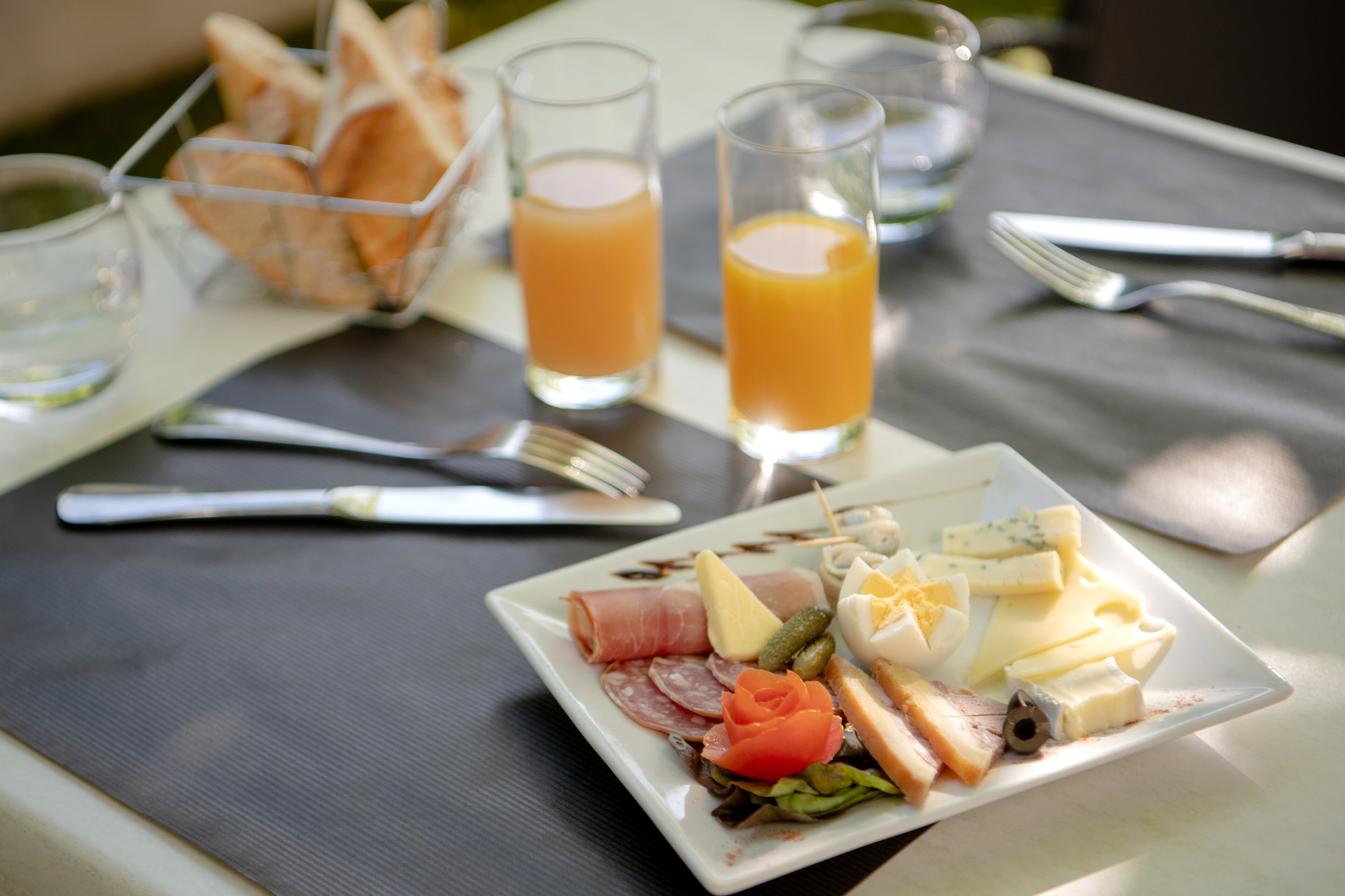 Una mesa dispuesta con un plato de quesos, carnes y verduras variados, junto con vasos de jugo de naranja y una cesta de pan.
