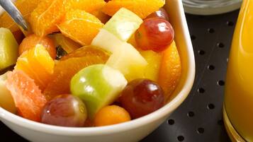 A bowl of mixed fruit salad with a spoon, placed on a black surface with a white bowl and a glass of orange juice nearby.