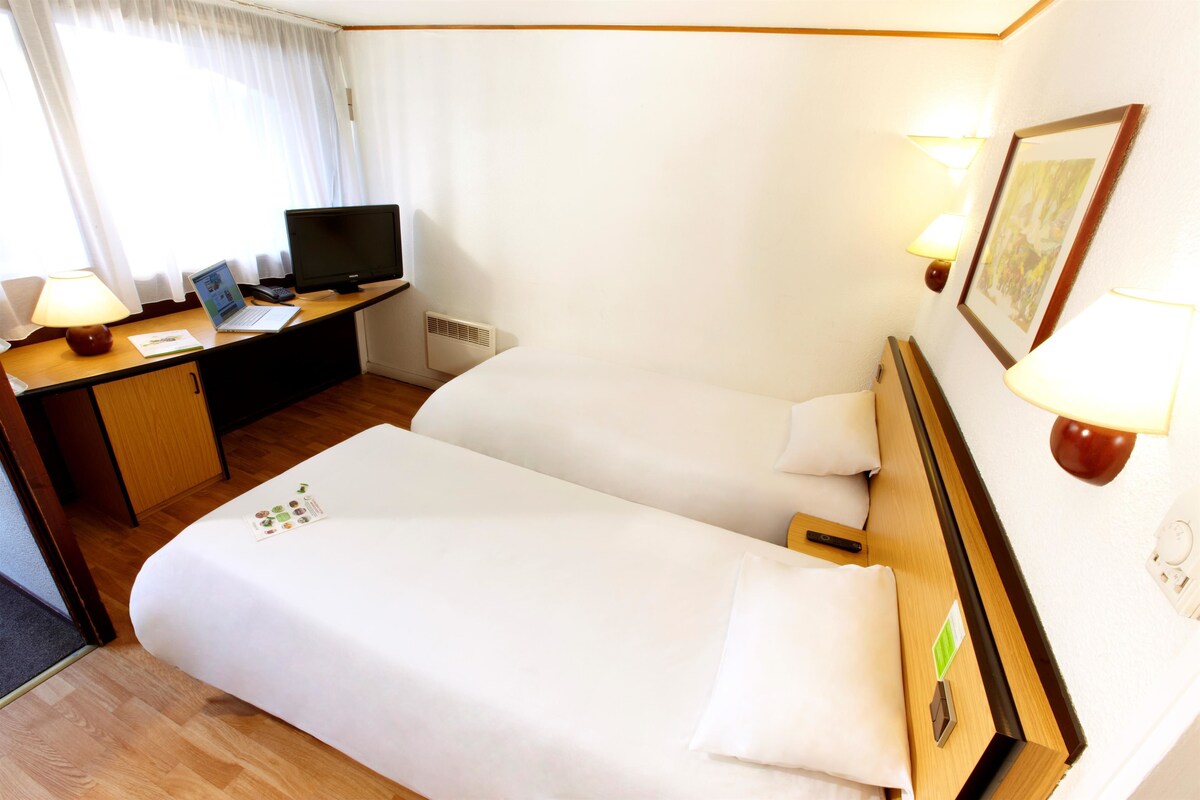 A hotel room with a bed, a desk with a laptop, a television, and a wall-mounted lamp.