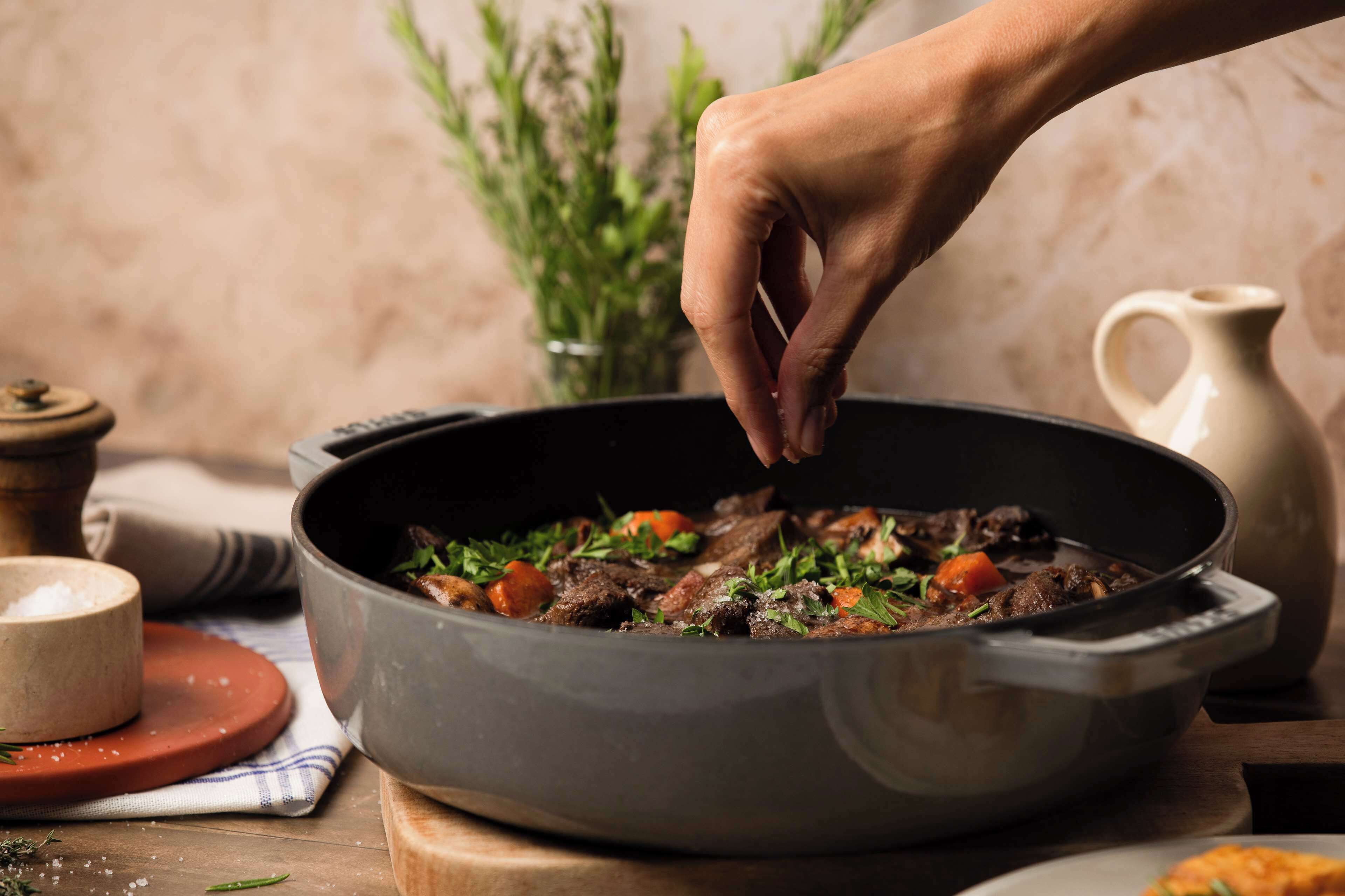 A hand stirring a stew in a pan on a wooden table.
