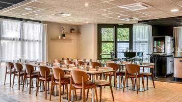 Un espace repas avec plusieurs tables et chaises, un coin cuisine équipé d’un four, et de grandes fenêtres laissant entrer la lumière naturelle.