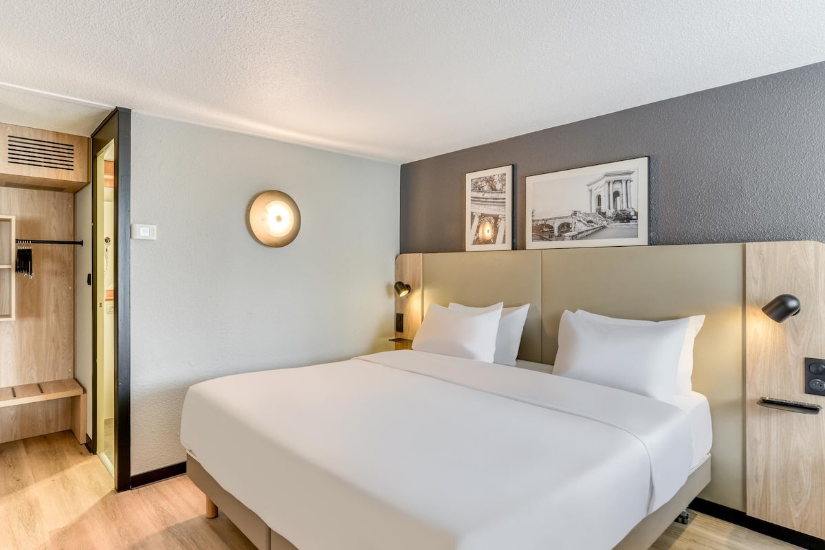 A hotel room with a large bed, a wardrobe, a bedside table, a wall-mounted light, and two framed pictures.