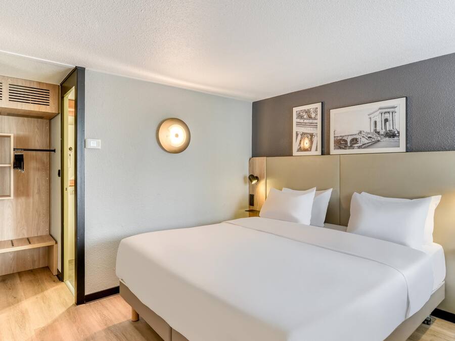 Une chambre d’hôtel avec un grand lit, une armoire, une table de chevet, une lampe fixée au mur et deux tableaux encadrés.