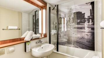 A modern bathroom with a large mirror, a white sink, a glass shower enclosure, and a view of a cityscape through the shower door.