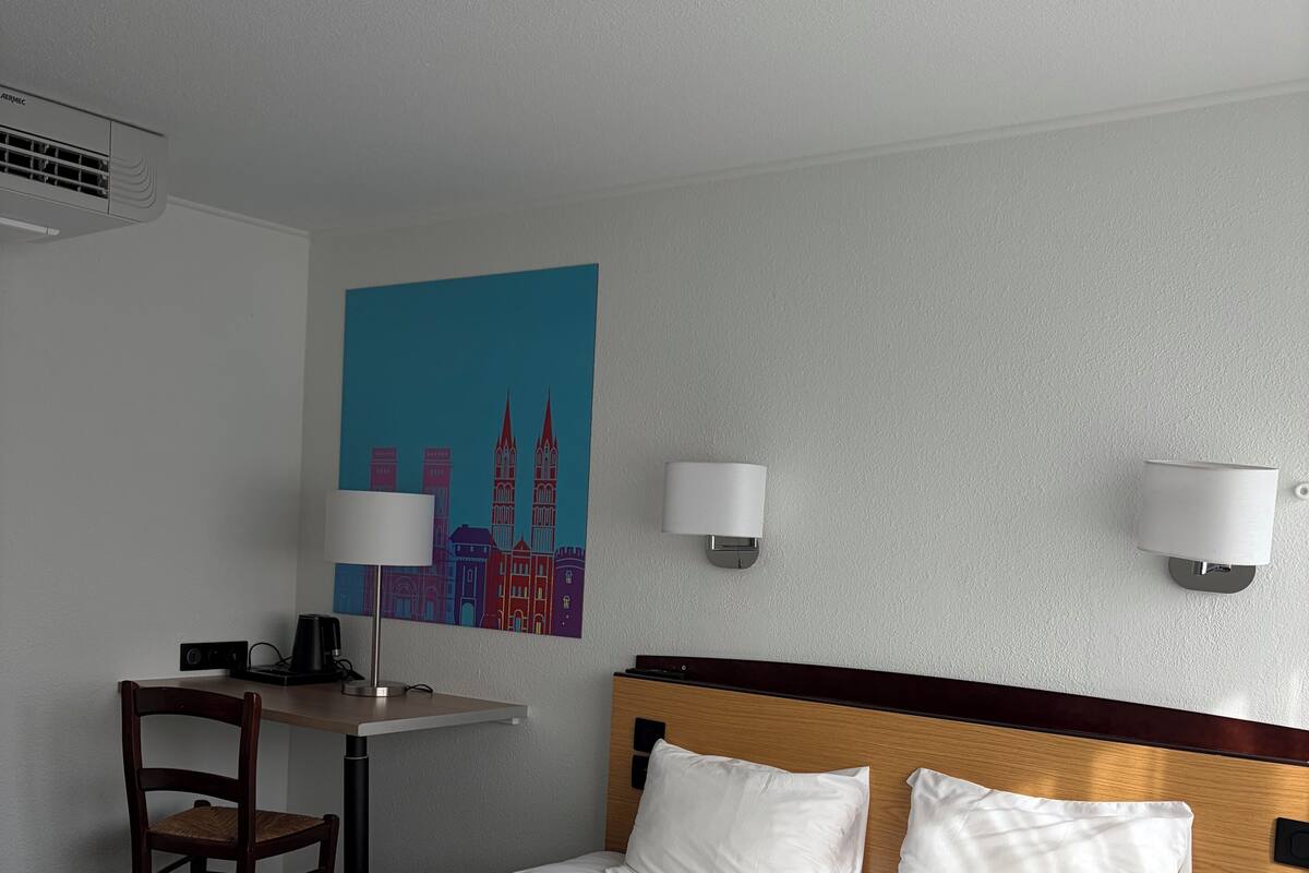 A hotel room with a bed, a desk, a chair, a wall-mounted lamp, and a painting of a cityscape.