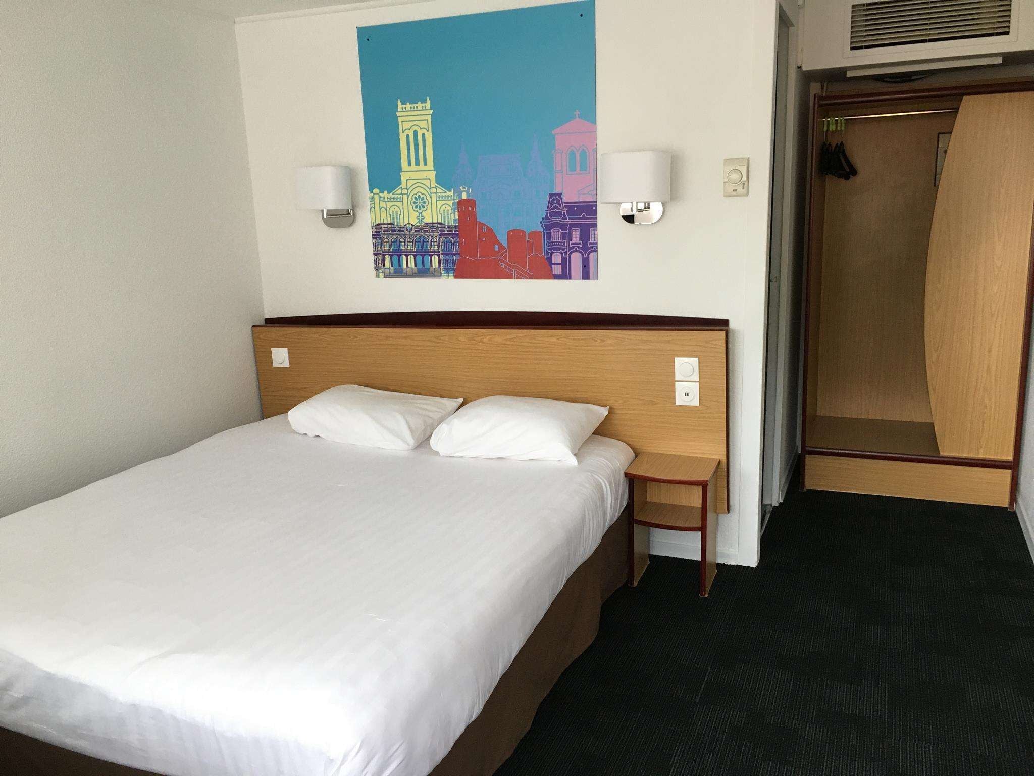 A hotel room with a bed, a nightstand, a wall-mounted lamp, and a door leading to a closet.