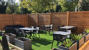 An outdoor patio area with wooden tables and chairs, surrounded by a wooden fence and green artificial grass.