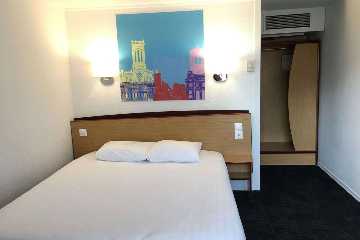 A hotel room with a bed, a wall-mounted artwork of a cityscape, a light switch, and a door leading to a closet.