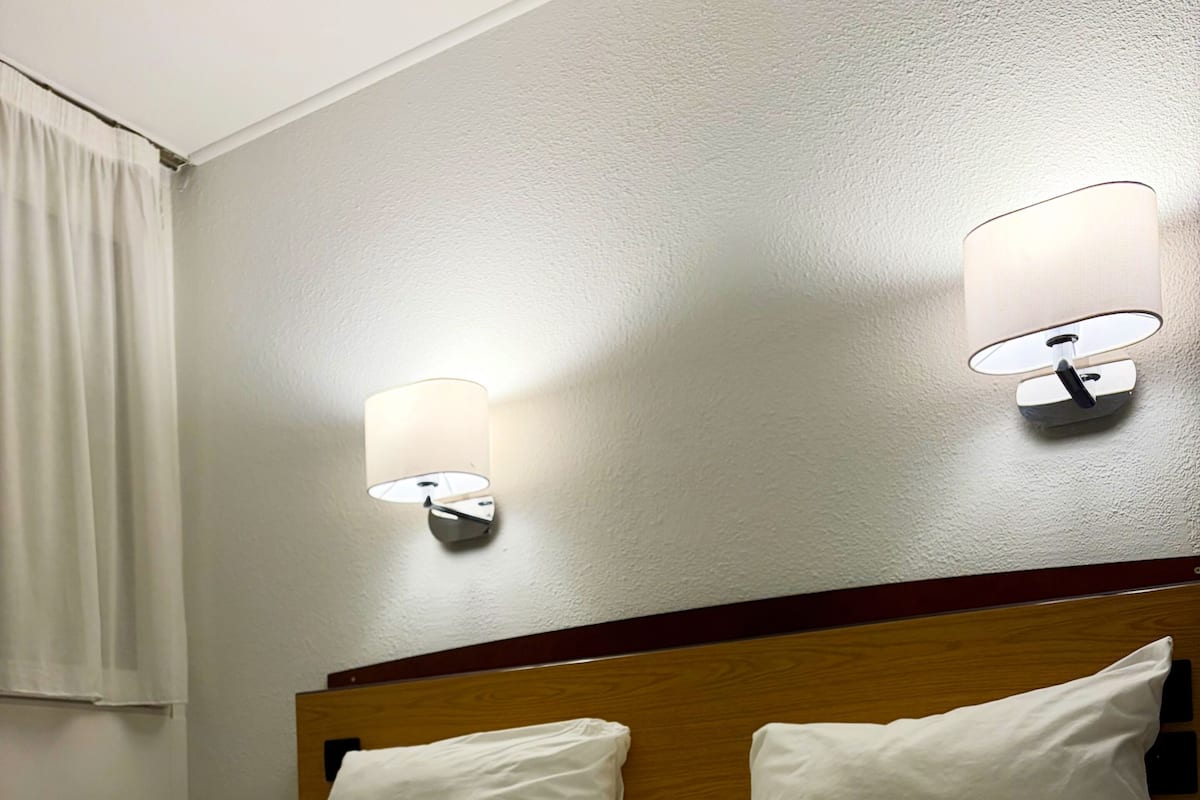 A hotel room with a neatly made bed, two wall-mounted lamps, and a wooden headboard.