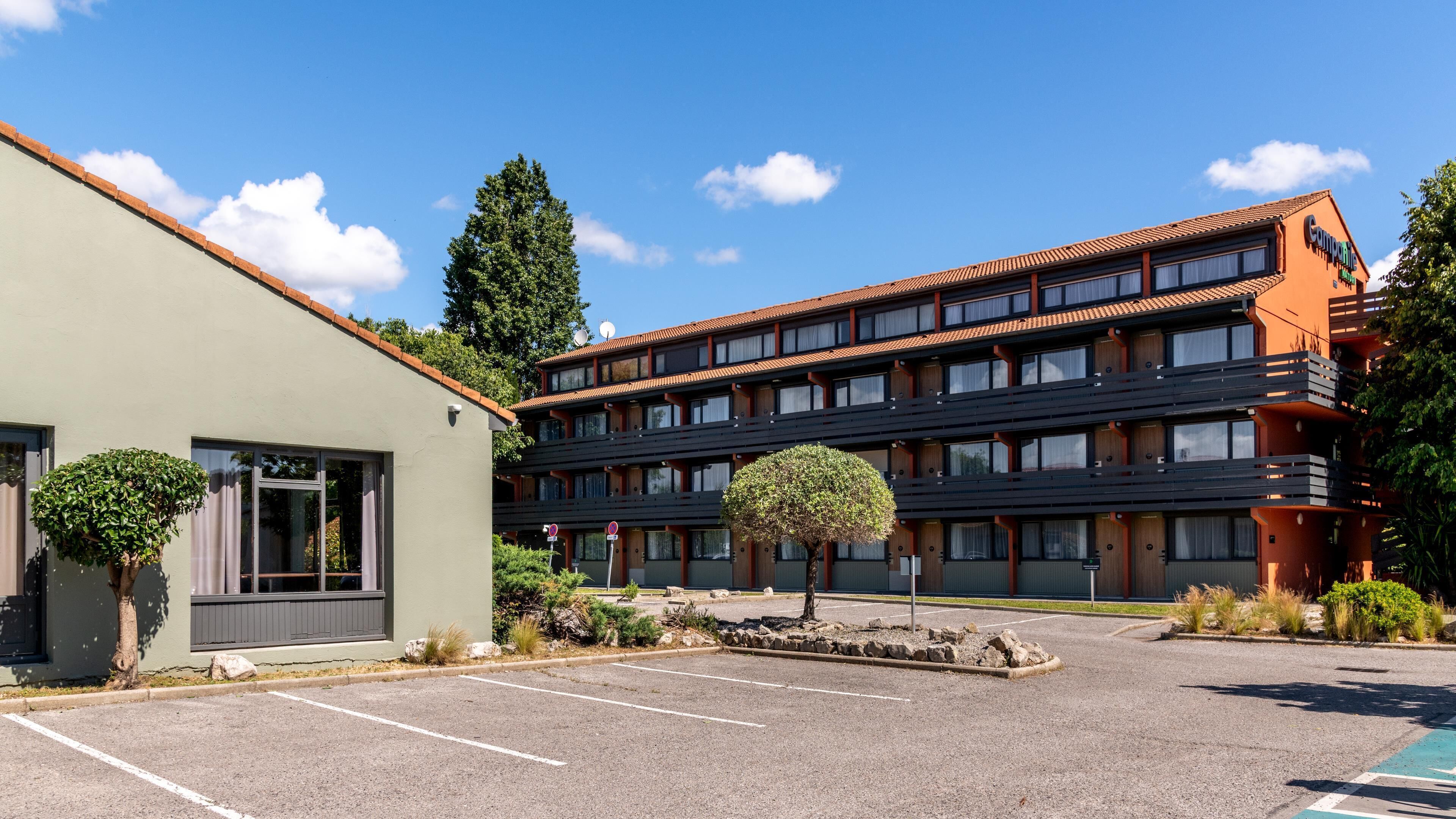 Un hôtel à plusieurs étages avec un parking, un petit arbre au premier plan et un ciel bleu dégagé.