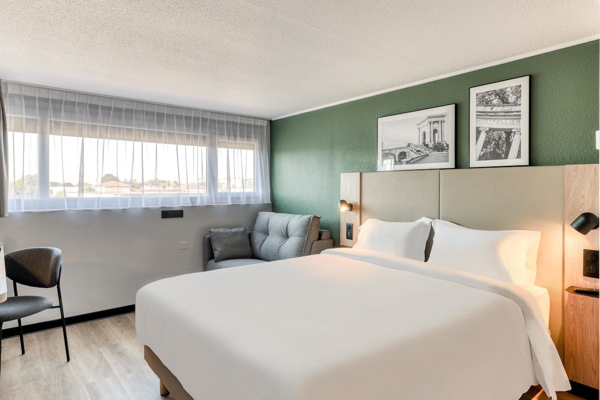 Een hotelkamer met een groot bed, een bureau met stoel, een raam met dunne gordijnen en twee ingelijste foto's aan de muur.