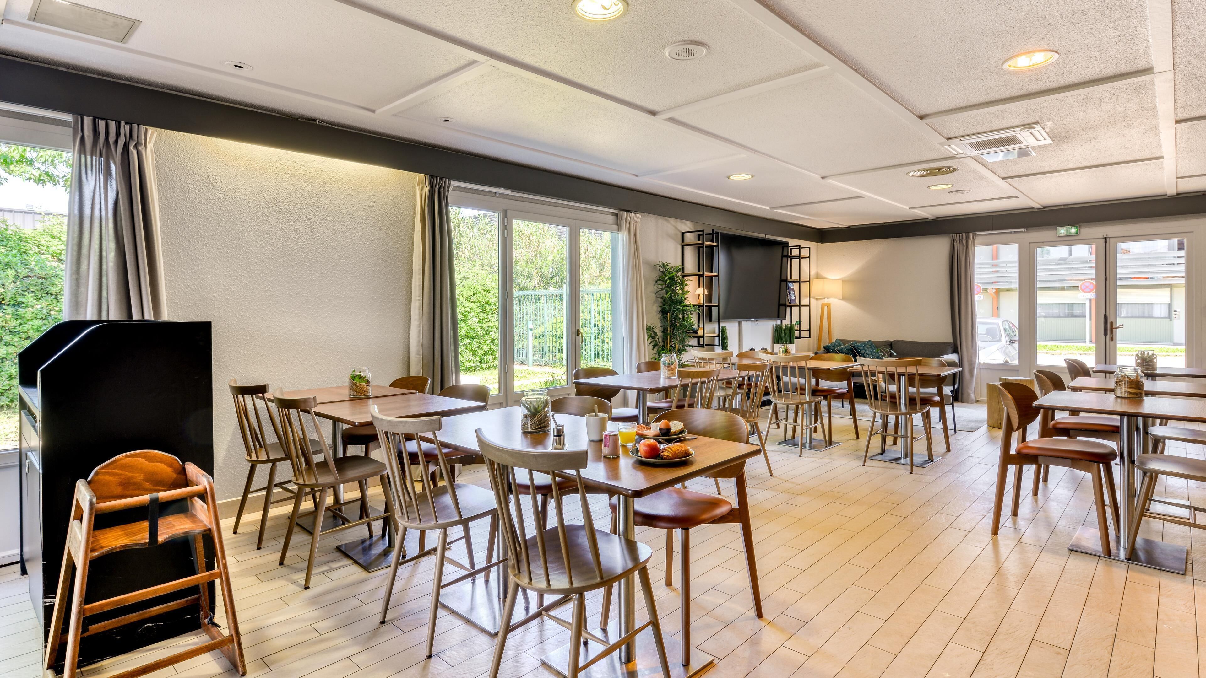 A spacious dining area with multiple tables and chairs, large windows offering views of greenery, and a modern ceiling design.