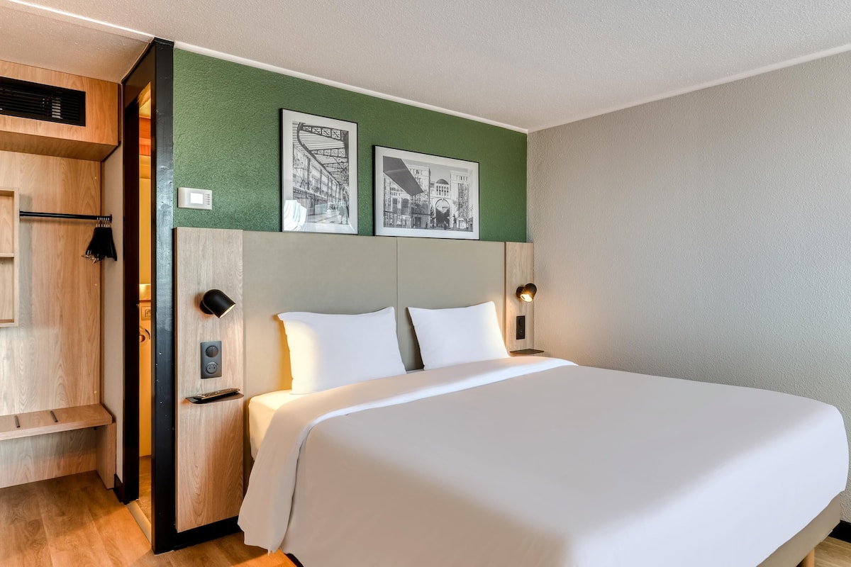 Een net opgemaakt bed in een hotelkamer met een groene accentmuur, een wandgemonteerde televisie en twee ingelijste foto's boven het bed.