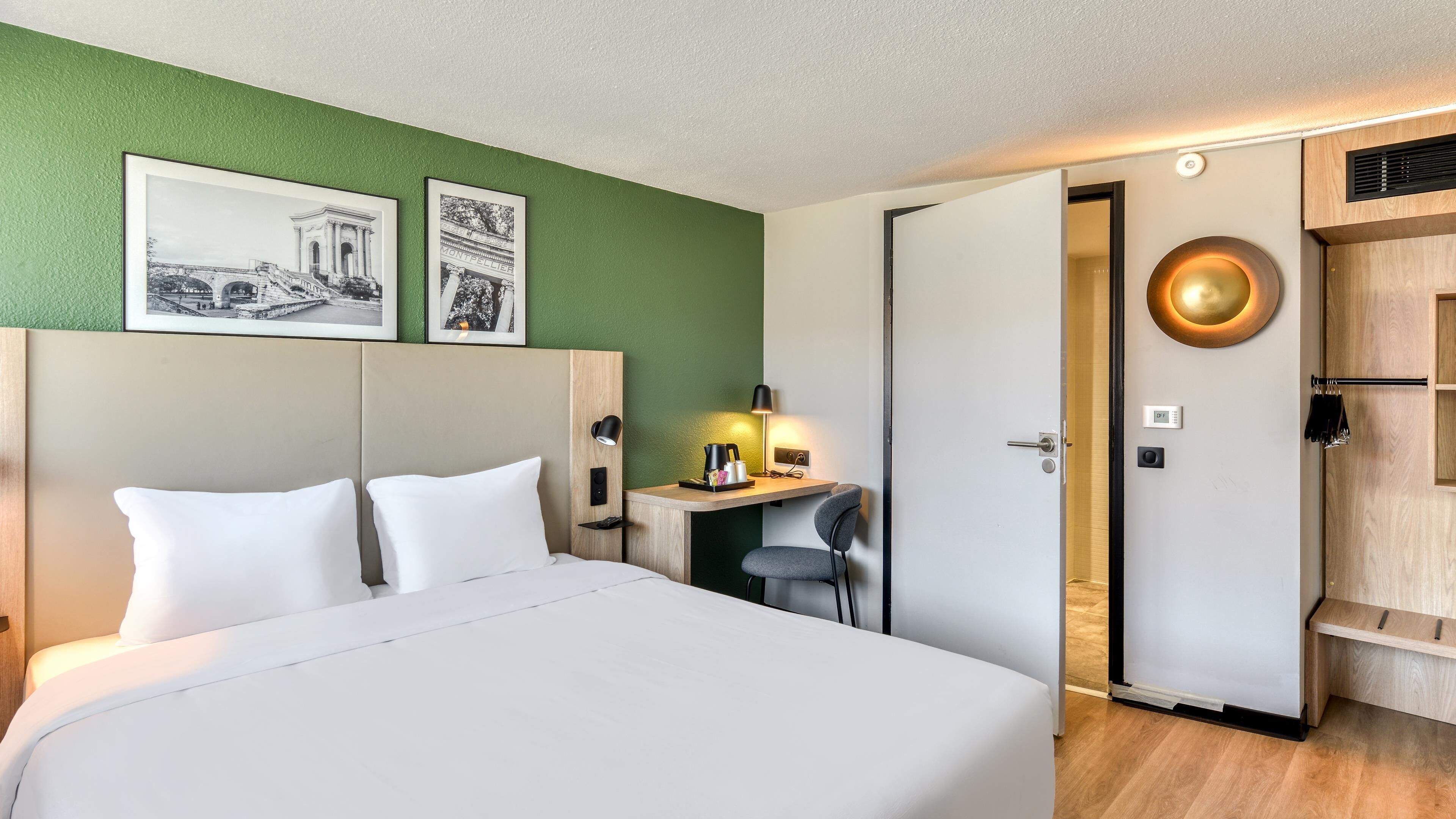 Habitación de hotel con cama, escritorio y silla. Hay dos cuadros en la pared, una lámpara en el escritorio y una puerta que da a otra habitación.