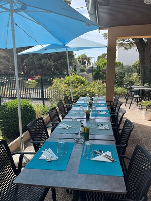 An outdoor dining area with tables set for service, surrounded by greenery and a fence.