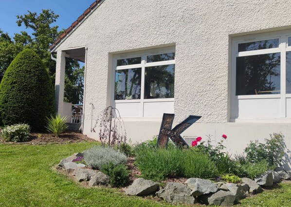 Un bâtiment blanc doté de grandes fenêtres, un jardin bien entretenu avec des rochers et des plantes, et une sculpture en forme de « K » bien visible au premier plan.