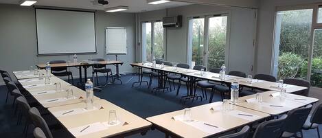 Un salón de conferencias con mesas largas, sillas, una pantalla de proyector y ventanas amplias.