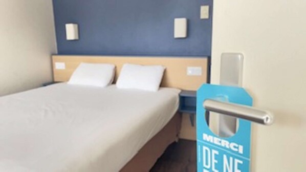 A hotel room with a bed, two pillows, a nightstand, and a blue wall with a sign that reads "MERCI DE NE".