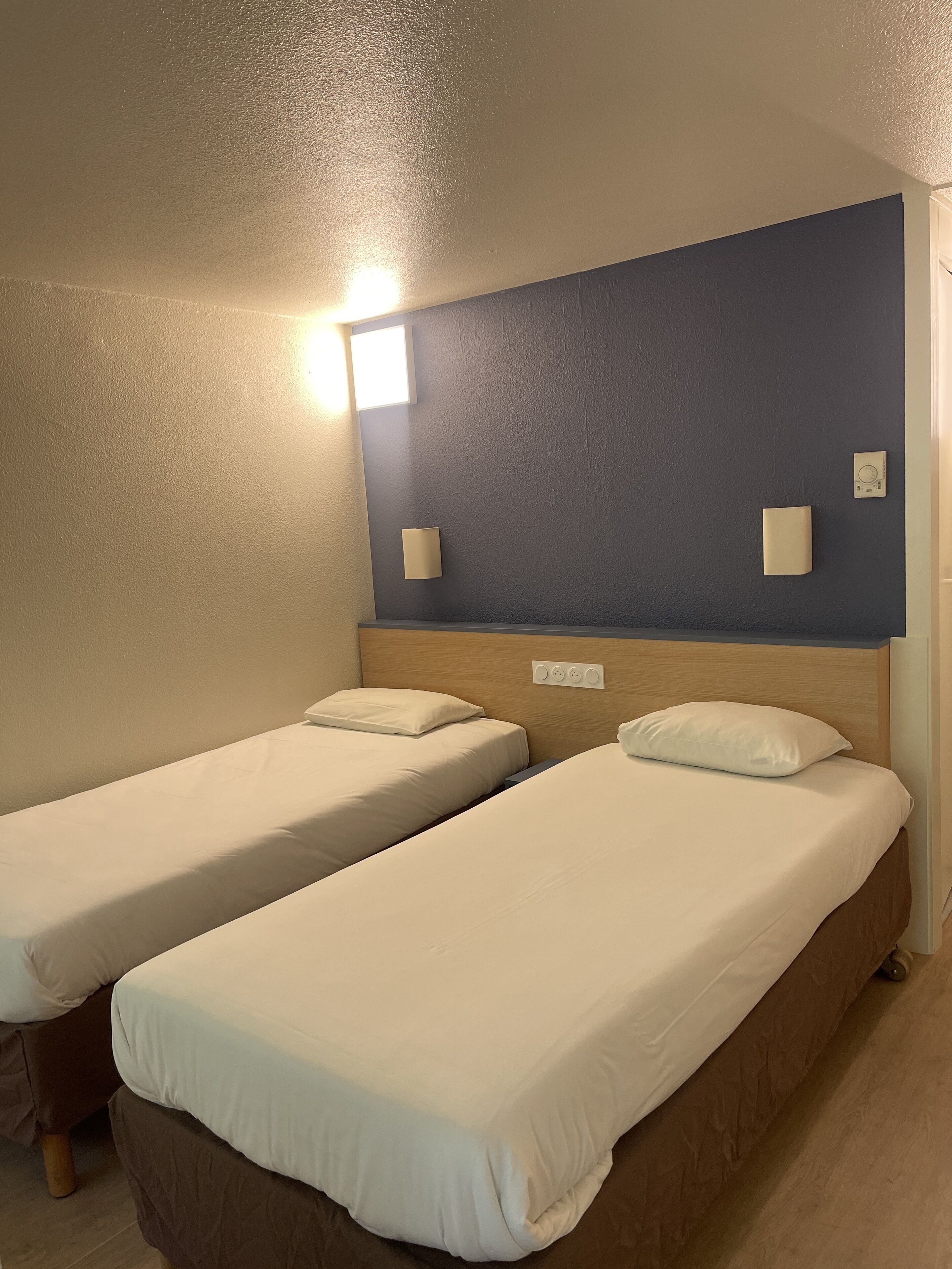 A room with two single beds, a wooden headboard, a wall-mounted light, and a power outlet.