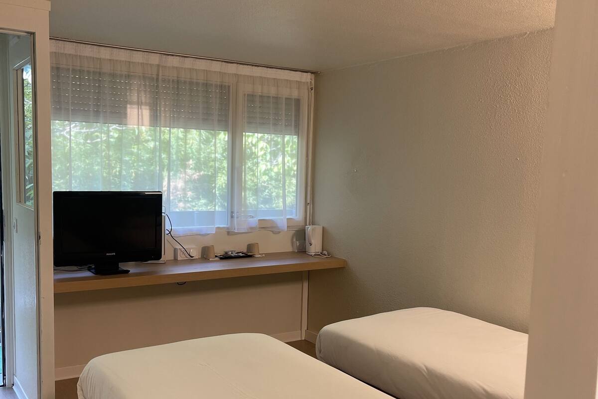 Ein Zimmer mit zwei Betten, einem Fernseher auf einem Holzregal und einem Fenster mit durchsichtigen Vorhängen.