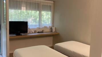 A room with two beds, a television on a wooden shelf, and a window with sheer curtains.