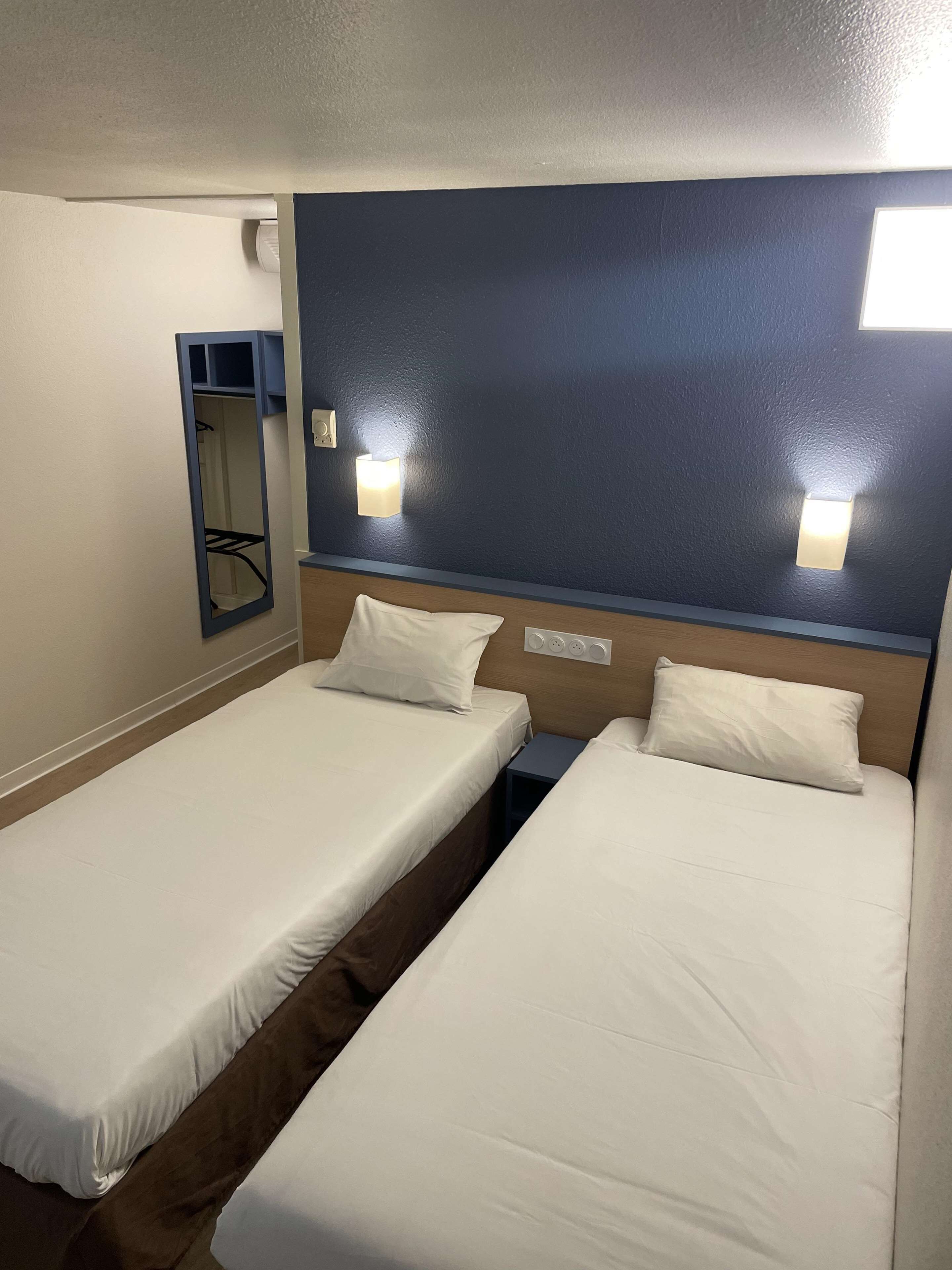 A hotel room with two single beds, a nightstand, a mirror, and wall-mounted lights.