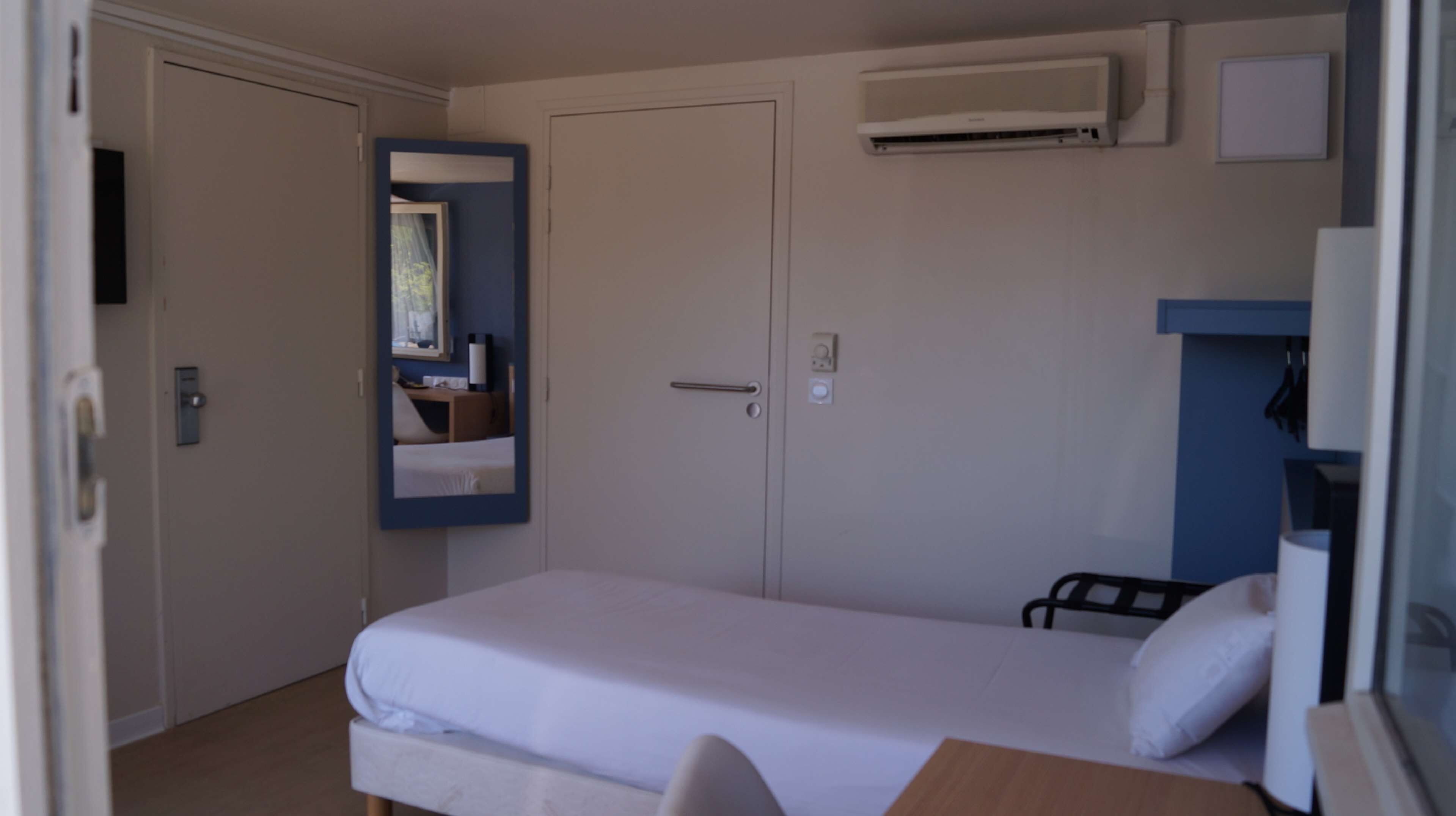 A compact bedroom with a single bed, a small desk, and a mirror.