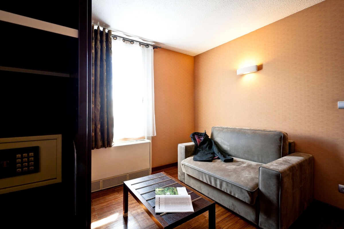 A room with a sofa, a coffee table with a magazine, a window with curtains, and a wall-mounted light fixture.