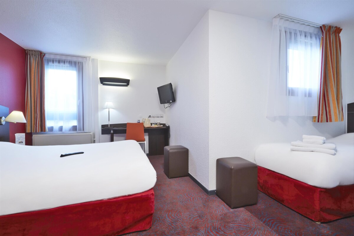 A hotel room with two beds, a desk, a chair, a TV, and a window with curtains.