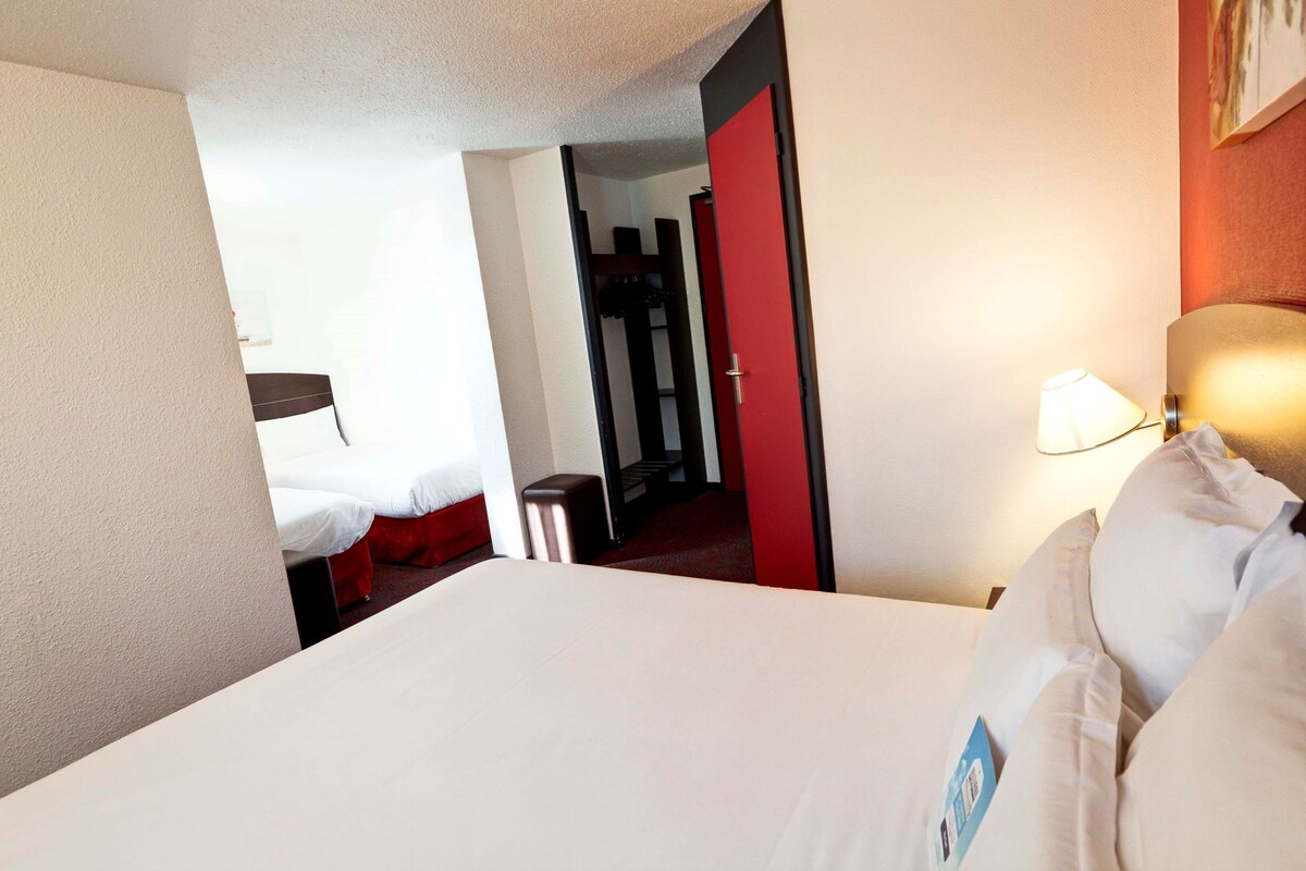 A hotel room with a bed, a bedside lamp, and a red door leading to a hallway.