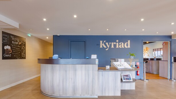 A reception desk with a blue wall and a door in the background. - Hôtel Kyriad Lyon Est (Saint-Bonnet-de-Mure)