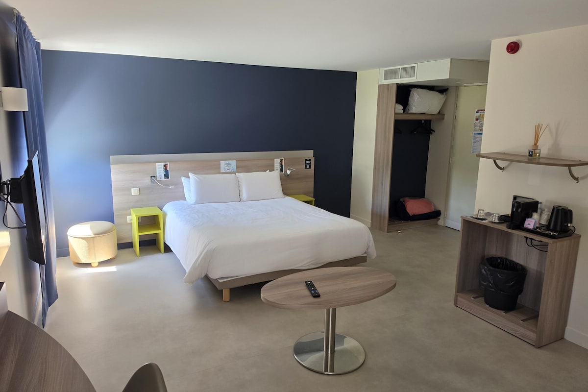 A hotel room with a bed, a desk, a chair, a small table, a trash bin, and a shelf with toiletries.