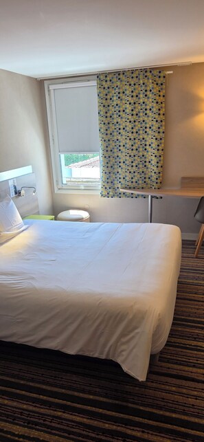 A hotel room with a bed, a desk, a chair, a window with a curtain, and a view of the outside.