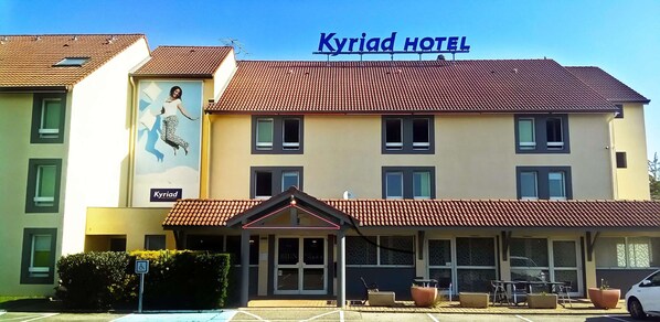 A hotel with a red-tiled roof, a sign reading "Kyriad Hotel," and a large mural on the side.