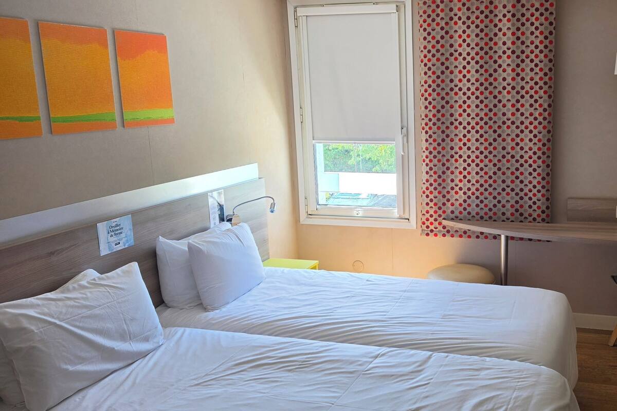 A hotel room with two beds, a desk, a chair, a TV, and a window with curtains.