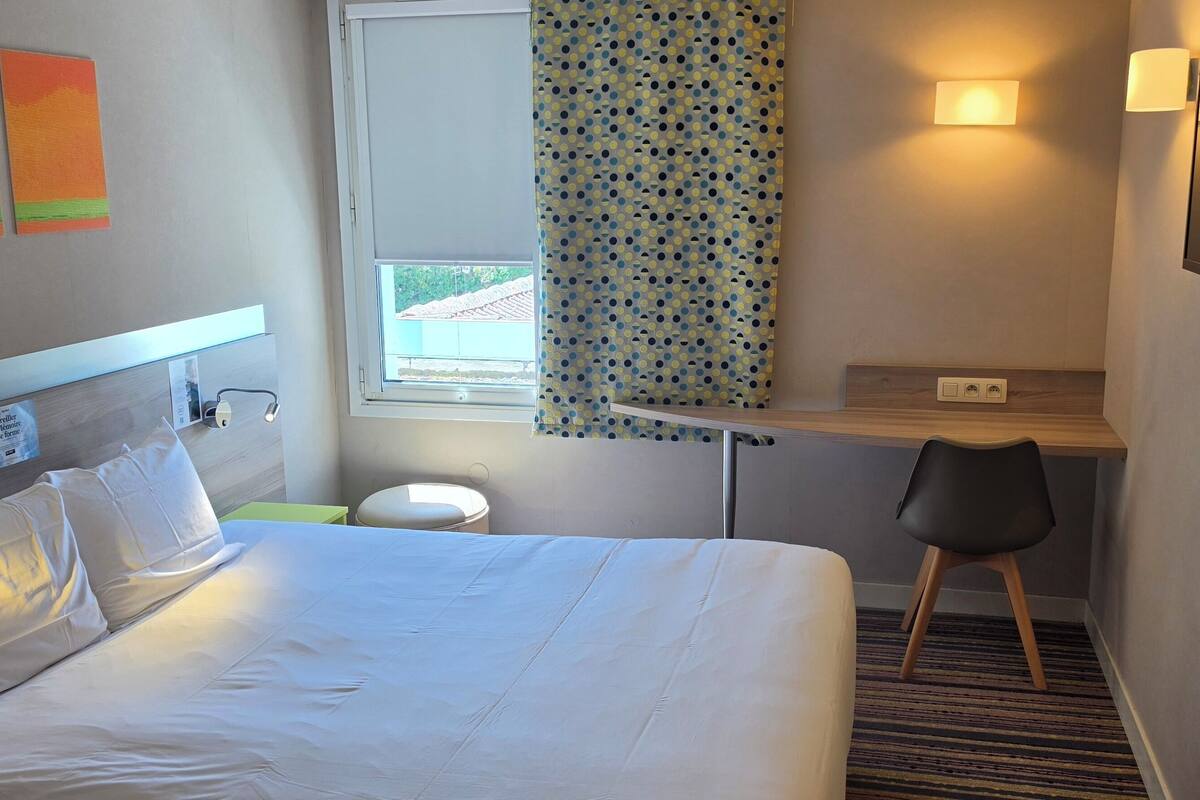 A hotel room with a bed, a desk, a chair, a window, and a wall-mounted TV.