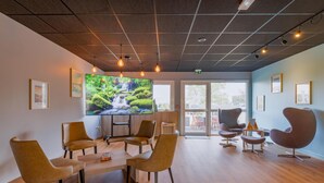 A modern meeting room with a large screen, comfortable seating, and a view through the glass door. - Hôtel Kyriad Lyon Est (Saint-Bonnet-de-Mure)