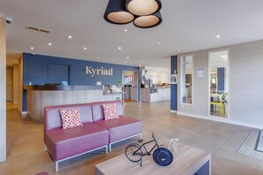 A modern hotel lobby with a reception desk, a sofa, a bicycle, and a kitchen area.