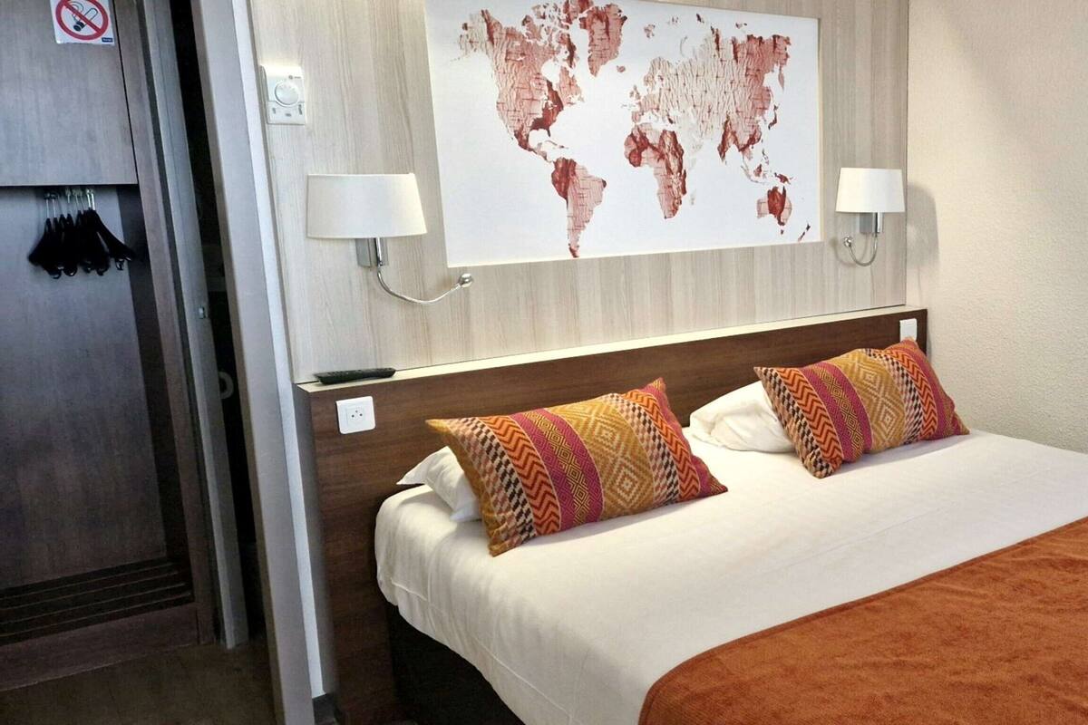 A hotel room with a large bed, two patterned pillows, a world map on the wall, and a brown throw blanket.