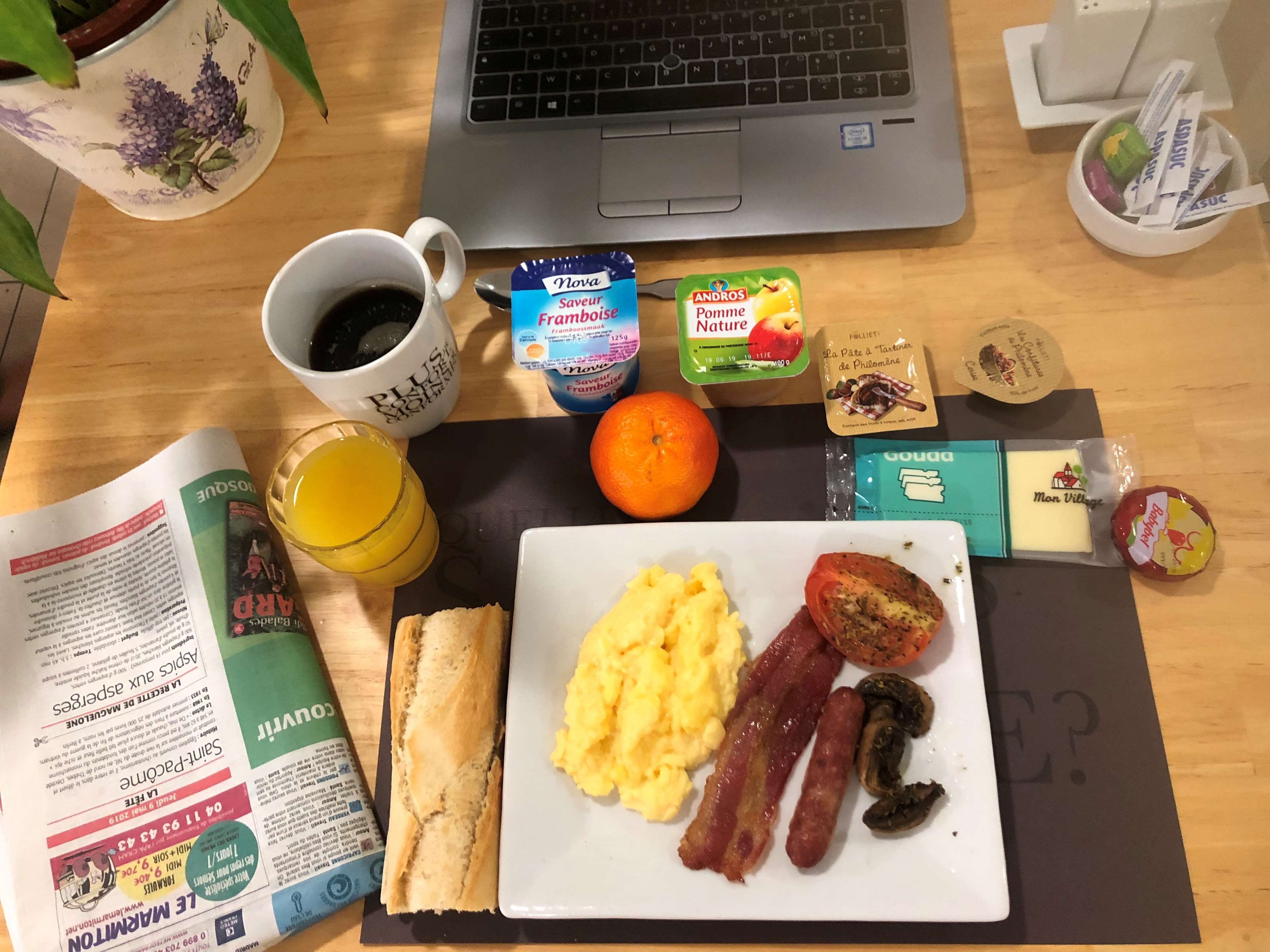 Een ontbijt uitgestald op een houten tafel, met een laptop, een koffiekop, een glas sinaasappelsap, een bord met roerei, spek, champignons, een baguette, een boek en diverse etenswaren.