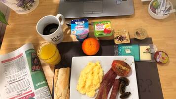 Een ontbijt uitgestald op een houten tafel, met een laptop, een koffiekop, een glas sinaasappelsap, een bord met roerei, spek, champignons, een baguette, een boek en diverse etenswaren.
