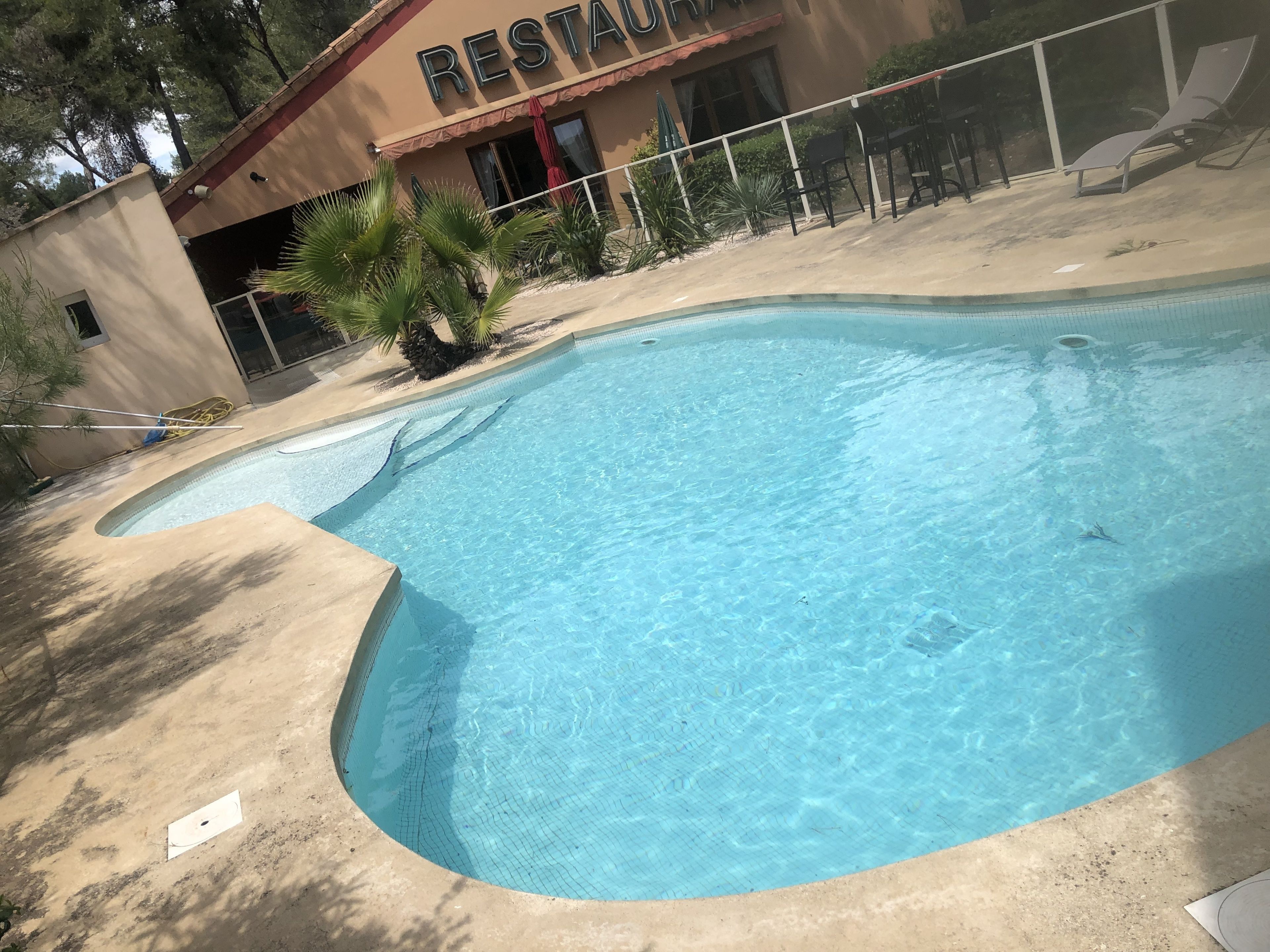 Une piscine à bord incurvé, un bâtiment abritant un restaurant en arrière-plan, et un espace extérieur aménagé avec des chaises et des parasols.